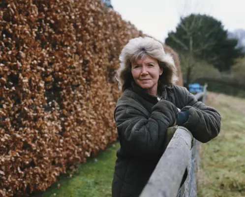 Jilly Cooper leaning on a fence in a field looking at the camera with a hedge behind her