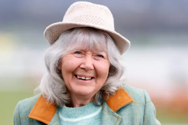 Close-up of Dame Jilly Cooper, wearing a plain hat and a light blue jacket and jumper, smiling.