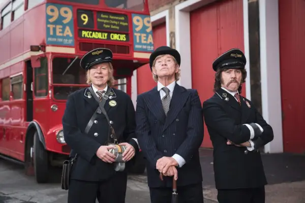 Steve Pemberton and Reece Shearsmith wear old-fashioned bus uniforms, while Robin Askwith stands between them in a suit. There is a red London bus, showing the number 9, behind them
