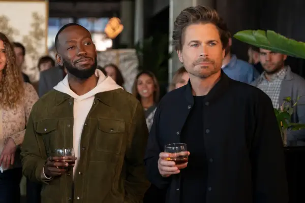 Peter and Ellis stand, holding glasses of alcohol at a function, looking at something to the right