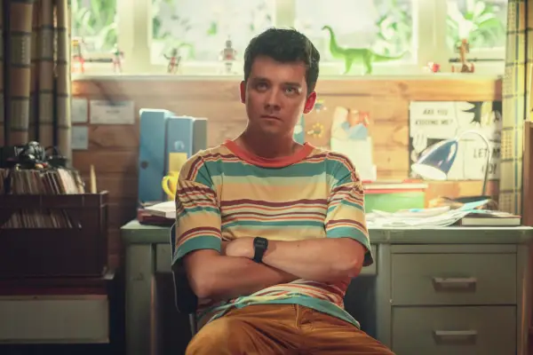 Asa Butterfield stars in Sex Education season 4. He is sitting down with his arms crossed. He is wearing a striped t-shirt.