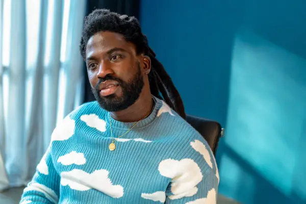 Adjani Salmon as Kwabena in Dreaming Whilst Black wearing a blue jumper with white clouds on it, staring off into the distance daydreaming.