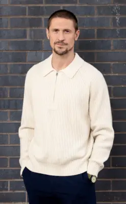 Jonathan Howard poses against a grey brick wall as Carl Webster in a promo shot for Coronation Street.