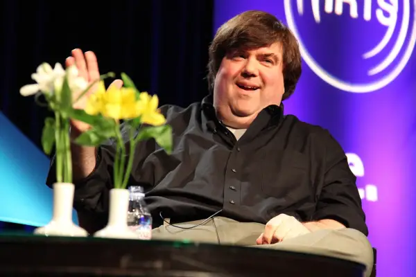 Dan Schneider sitting on stage at a table with yellow flowers on it. There is a purple board in the background.
