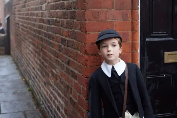 Dainton Anderson as child Archie in Archie hiding round a street corner wearing a school uniform.