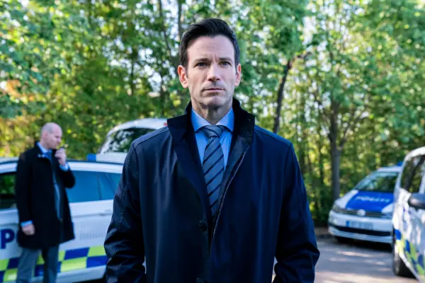 Colin Donnell as Brian Lange standing in the middle of a forest wearing a suit and coat, which he has his hands in.