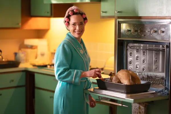 Laura Main as Shelagh Turner standing in the kitchen in her dressing gown and rollers next to a turkey in the oven