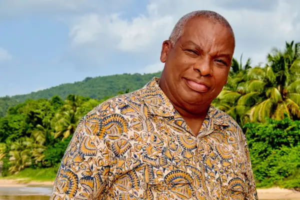 Commissioner Selwyn (Don Warrington) stands on the beach, wearing a colourful patterned shirt