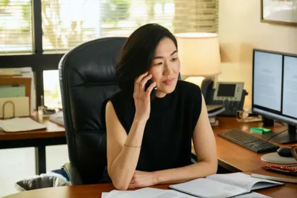 Jackie Chung as Laurel, sat in her office at her desk, on the phone