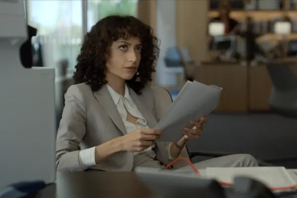 Rosaline Elbay as Nora in The Diplomat, sat in a chair and looking at a document.