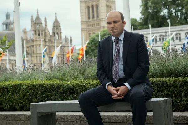 Rory Kinnear as Nicol Trowbridge in The Diplomat, sat on a bench with the houses of parliament behind him.