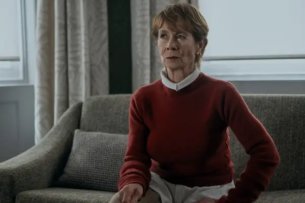 Celia Imrie as Margaret Roylin in The Diplomat, wearing a red jumper and sat on a sofa.
