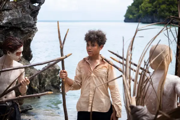 Winston Sawyers as Ralph in Lord of the Flies. He is stood by the sea holding a stick and two others and pointing sticks at him.