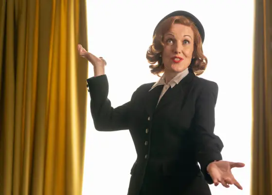 Kacey Ainsworth as Cathy Keating in Grantchester. She is wearing a uniform and is stood in front of two golden curtains, posing with her hands.
