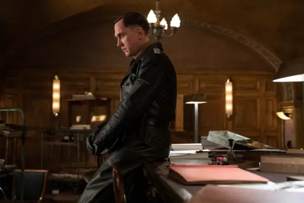 Lars Eidinger as Sergeant Major Reinhold von Rumpel in All the Light We Cannot See perched on the side of a desk.