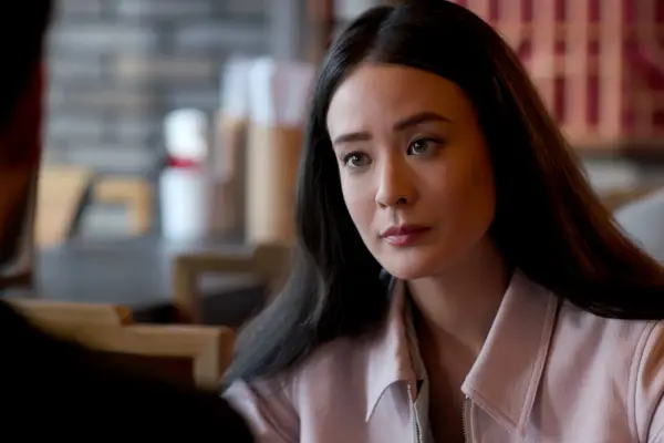 Highdee Kuan as Alexis in The Brothers Sun sitting at a restaurant wearing a pink jacket.