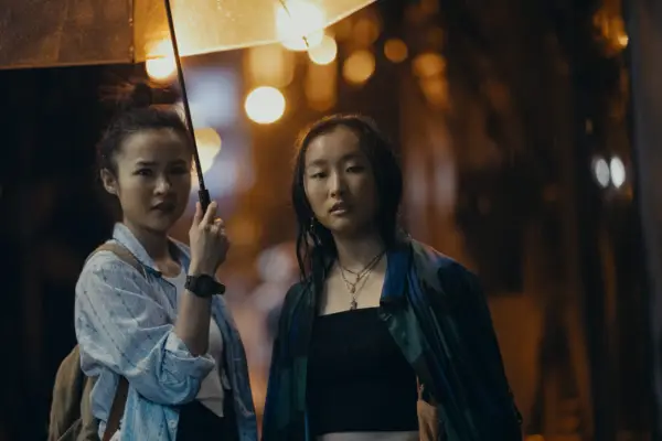 Bonde Sham as Charly holding a yellow umbrella over Ji-young Yoo as Mercy as they look down the street at something.