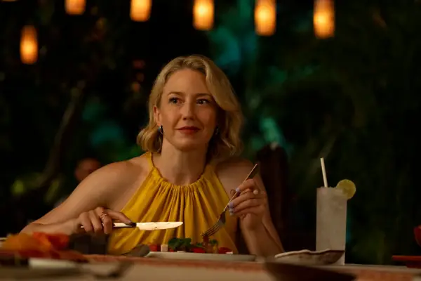 Carrie Coon wearing a yellow cut-off top as Laurie Duffy seated eating at a table in The White Lotus season 3.