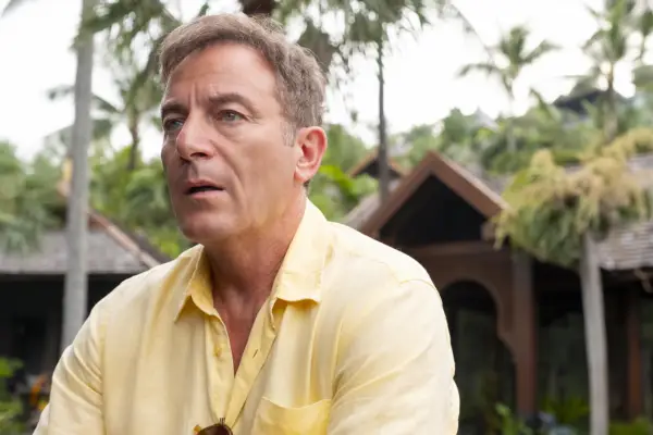 Jason Isaacs sat in an exterior setting in Thailand in a yellow shirt in close-up looking anxious as Timothy Ratliff in The White Lotus season 3.