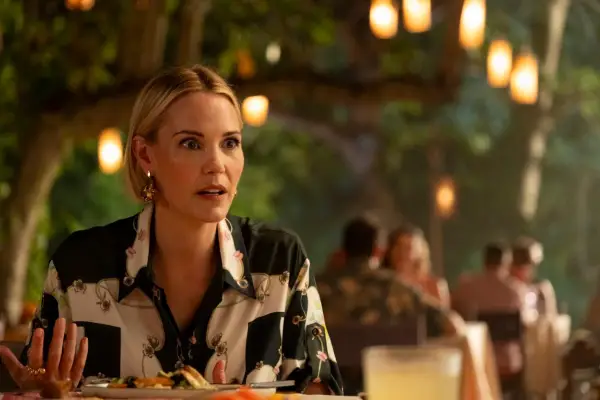 Leslie Bibb as Kate Bohr sat at a table in a white and black patterned shirt looking surprised in The White Lotus.