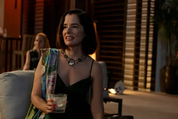 Parker Posey stands in a patterned dress holding a drink at am evening party as Victoria Ratliff in The White Lotus season 3.