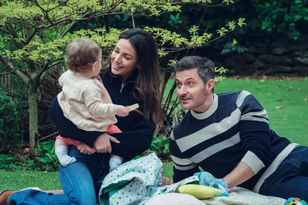 Maya kneeling on the grass holding her baby with Joe lying next to her