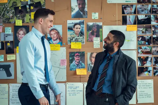 Two detectives standing next to each other in front of an evidence board, laughing