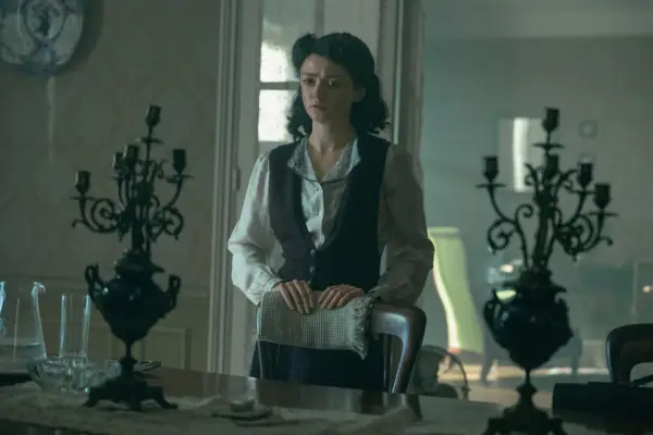 Maisie Williams as Catherine Dior wearing a shirt and dress, holding on to the top of a dining chair while standing.