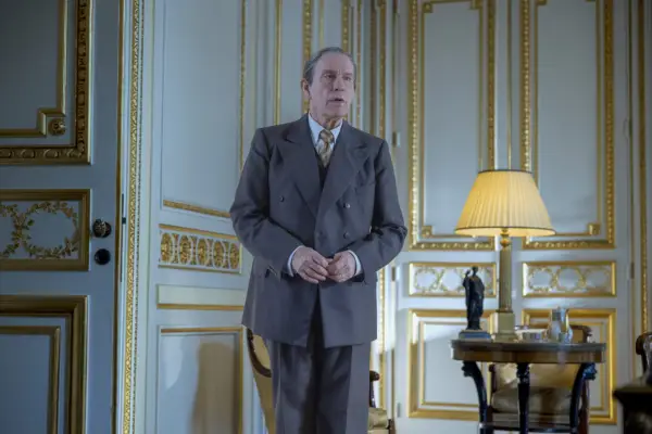 John Malkovich as Lucien Lelong in The New Look standing in a grey suit in a gilded white room with his hands held in front of him.