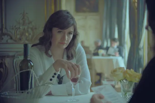 Emily Mortimer as Elsa Lombardi in The New Look holding on to a glass of wine while sat at a table.