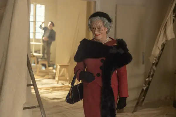 Glenn Close as Carmel Snow in The New Look wearing a matching red suit and black accessories, smiling at someone.
