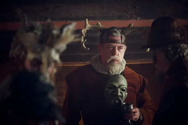 Sean Gilder as Sir Thomas Compton in Mary & George wearing a red suit and antlers on his head while holding a mask.