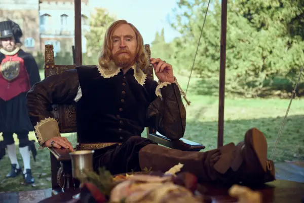 Tony Curran as King James in Mary & George sitting at a banquet table filled with food, resting his elbow and looking out.