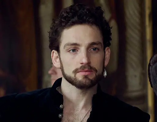 Niamh Algar wearing a burgundy dress and white collar, sitting on a chair with her arm propped up on a wooden table.