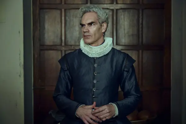 Angus Wright standing against a wood panelled wall wearing a blue suit and white collar, looking to his side.