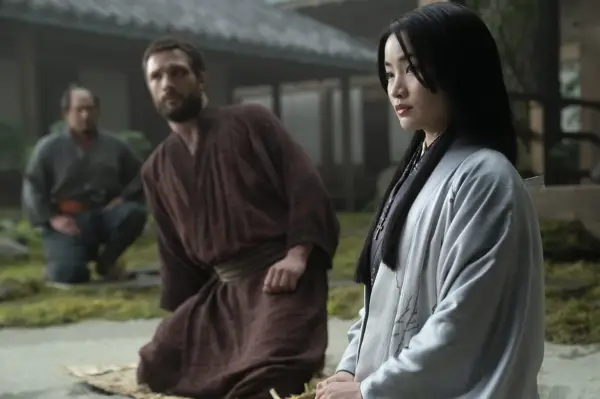 Cosmo Jarvis and Anna Sawai both wearing robes, sitting down on the floor outside looking at something.