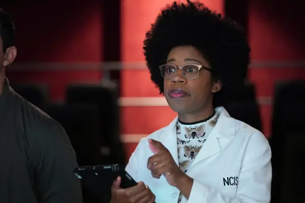 A woman in an NCIS-branded lab coat holds a tablet and gestures towards someone as if making a point