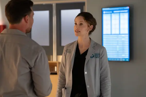 Sarah Ramos as Dr. Caitlin Lenox in Chicago Med season 11 standing in a dimly lit hospital ward, wearing her scrubs and lab coat.