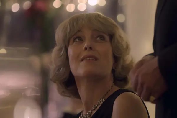 Olivia Williams portrays Camilla in The Crown; she is seated and craning her neck to look at somebody standing behind her