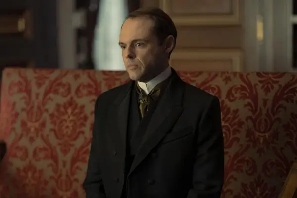 Christopher Denham as Robert McNeil in The Gilded Age sitting on a sofa.