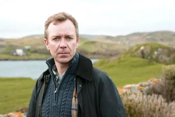 Steven Robertson plays Sandy Wilson, standing on a grassy bank, with the sea in the background, looking directly into the camera