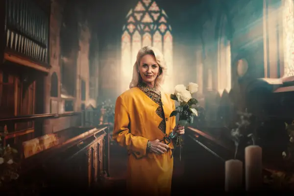 Tamsin Outhwaite as Stella Harper. stood in the middle of an aisle in a church, pews and a big stained glass window behind her, looking at the camera while holding white flowers, wearing a yellow coat