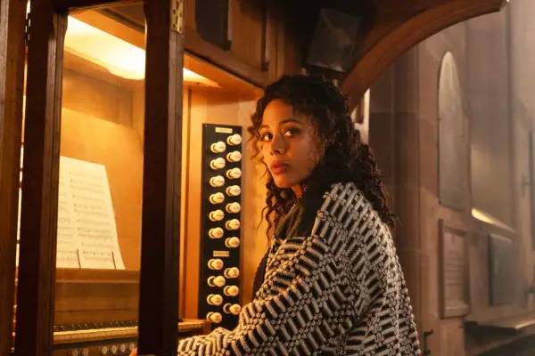 Nina Toussaint-White as Jane Thwaite, sat playing the organ while looking to her left, wearing a black and white patterned cardigan