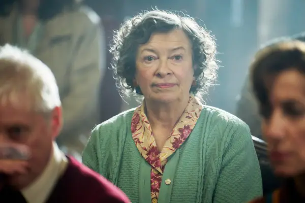 Marion Bailey as Kath Sharman, sat in a church pew, next to other people, wearing a green cardigan and floral blouse