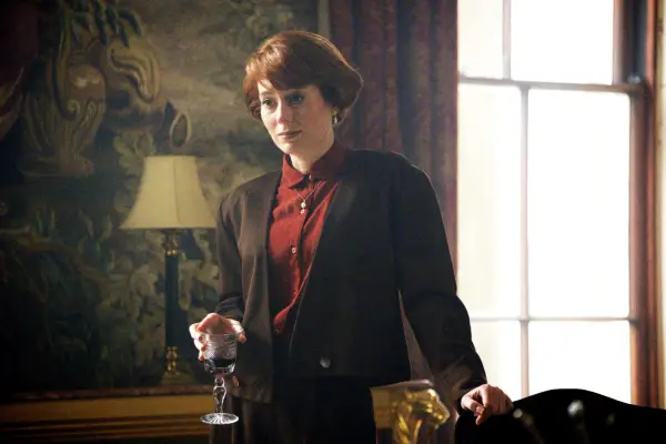 Meghan Treadway as Honor de Floures, stood in a living room, wearing a suit, holding a glass of alcohol