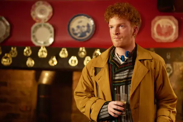 Alexander Delamain as Alex de Floures, standing in a pub, holding a glass of drink