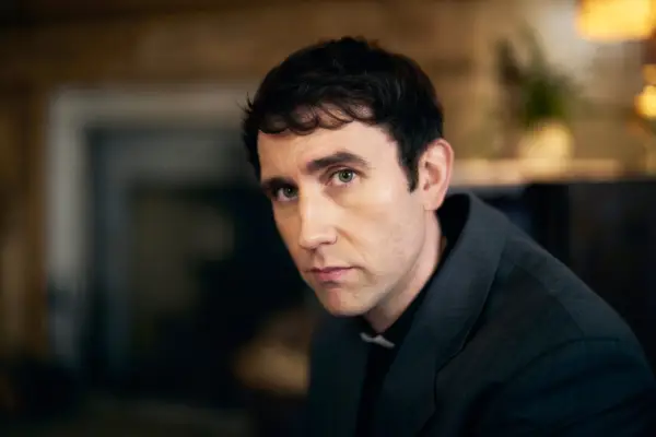 Matthew Lewis as Canon Daniel Clement, sat down in a room, dressed in black, looking directly at the camera