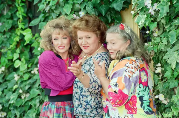 Three women from Keeping Up Appearances — Hyacinth Bucket stands in the centre with a confident smile, flanked by her sisters Rose and Daisy, all dressed in bright 1990s outfits, posing cheerfully in front of a wall covered with green ivy and flowers.