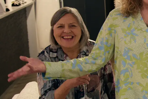 Nicola Cavendish as Connie. She is holding a flute of alcohol and smiling.