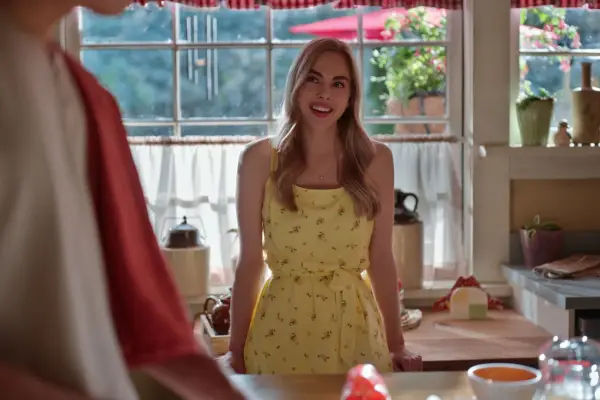 Sarah Dugdale as Lizzie wearing a yellow dress, smiling ahead as she stands in a kitchen.